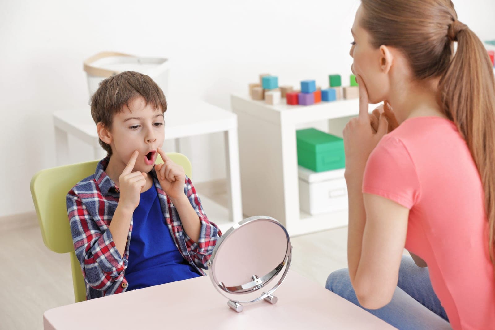 A speech-language pathologist and a young child practice speech sounds together using a hand mirror.