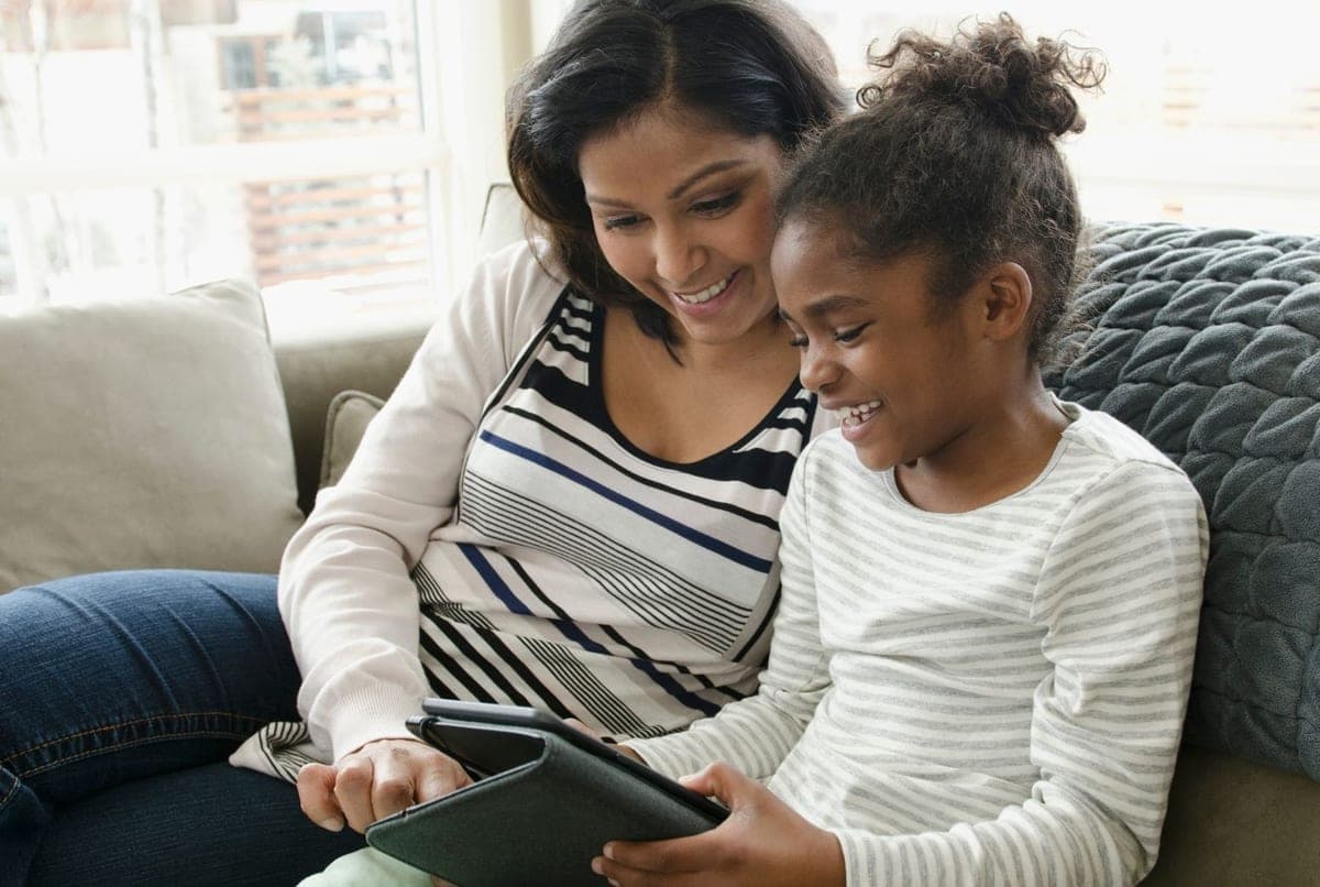 A parent and child sitting on a sofa, smiling at a tablet screen together